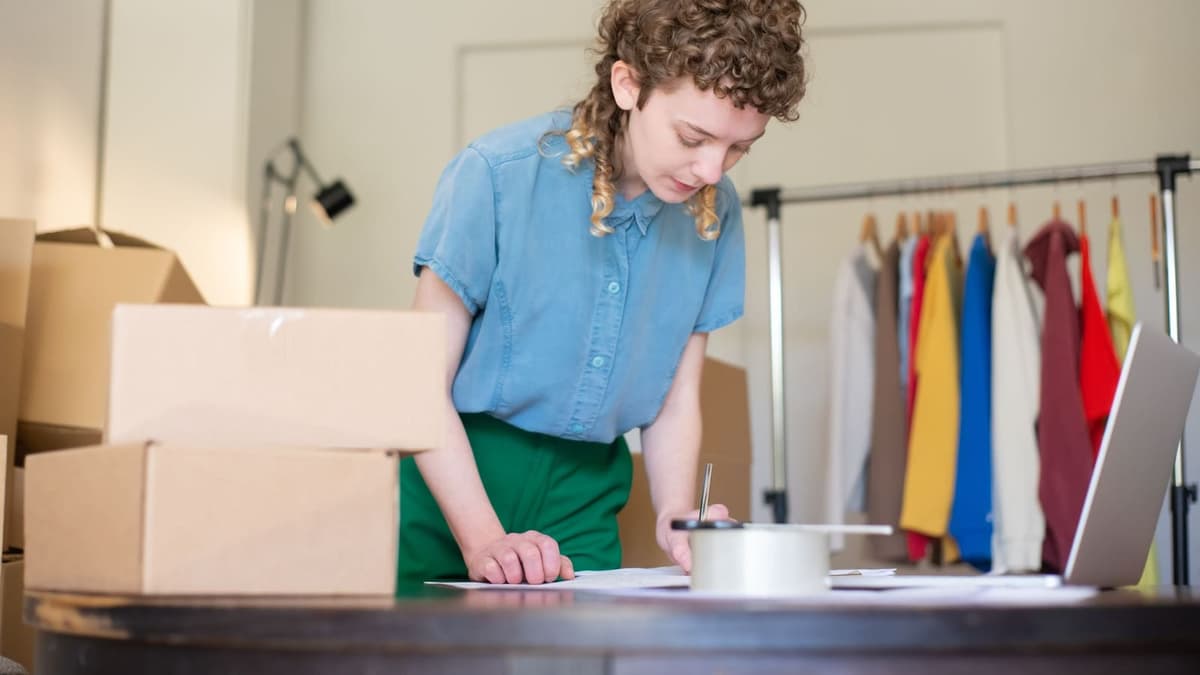 Lojista embalando pedidos em caixas de papelão ao lado de uma arara de roupas